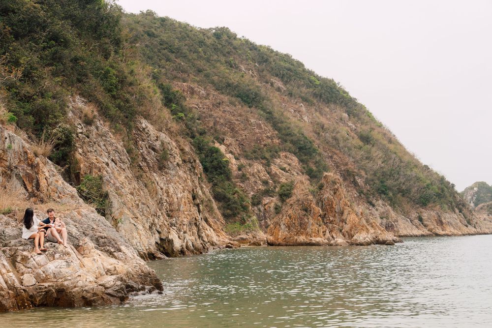 Two people sitting on rocky coastal cliffs beside calm green waters with a lush forested hillside in the background.