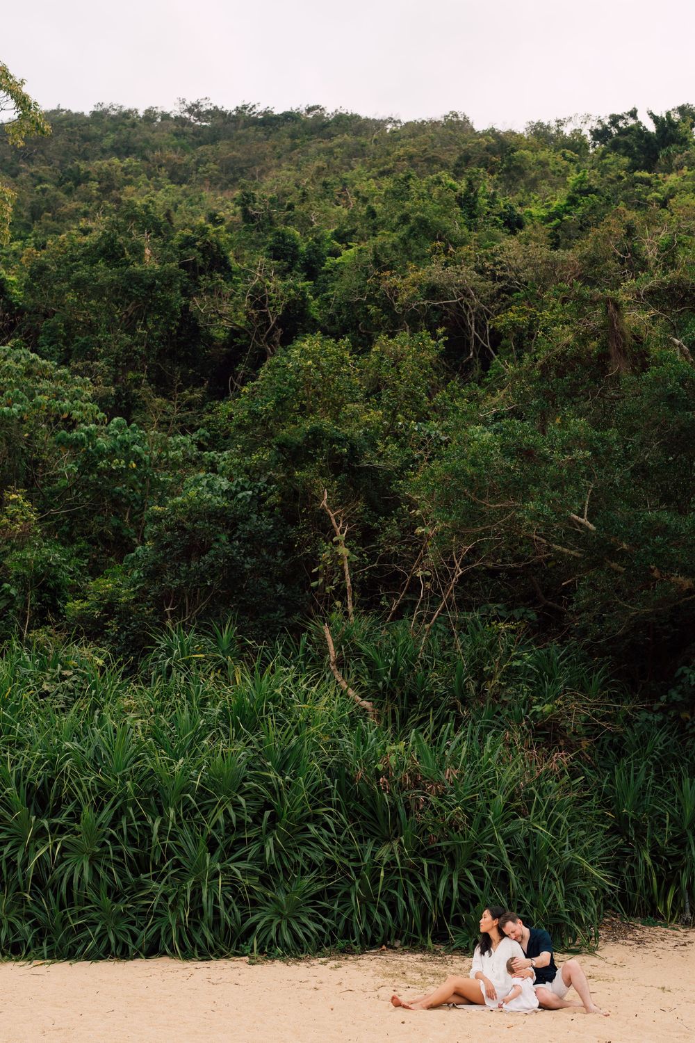 Couple sitting together on sandy beach with lush green tropical forest and hillside in background.