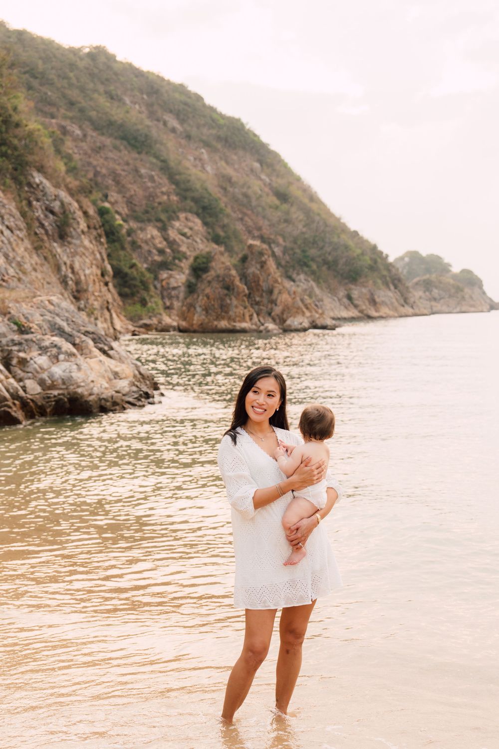 Smiling Asian mother in white dress holds baby while standing in shallow ocean water near rocky cliffs.