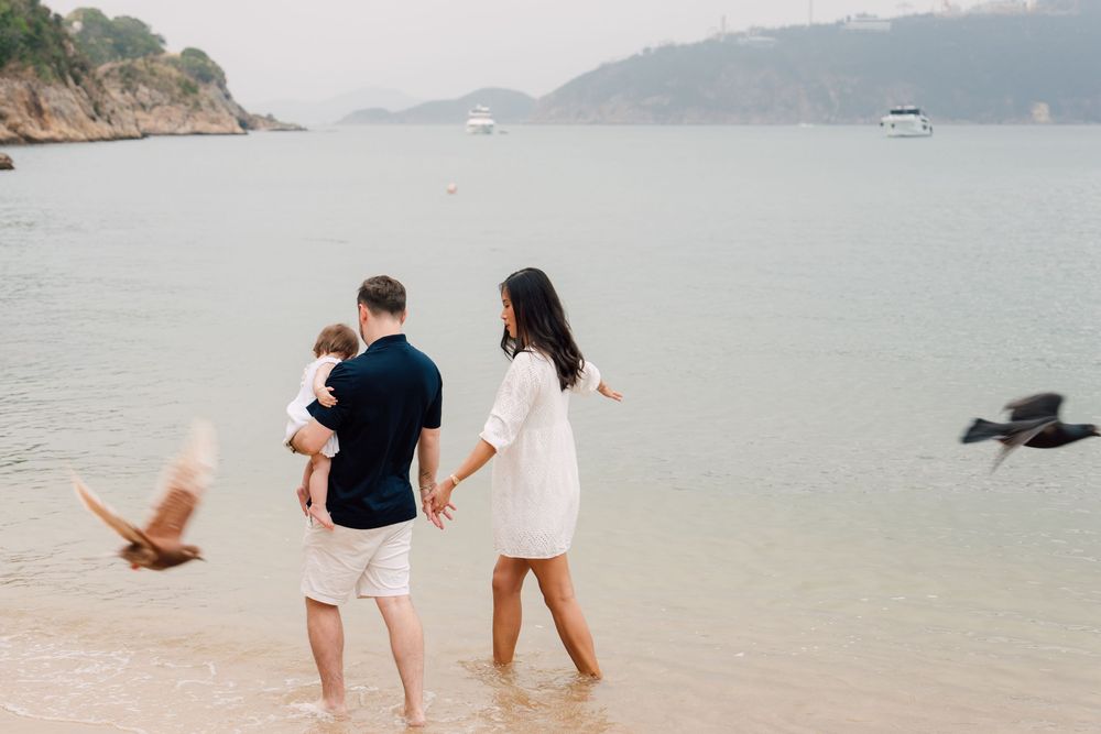 Family of three standing in shallow ocean water on a tropical beach, watching birds fly by on a misty day.
