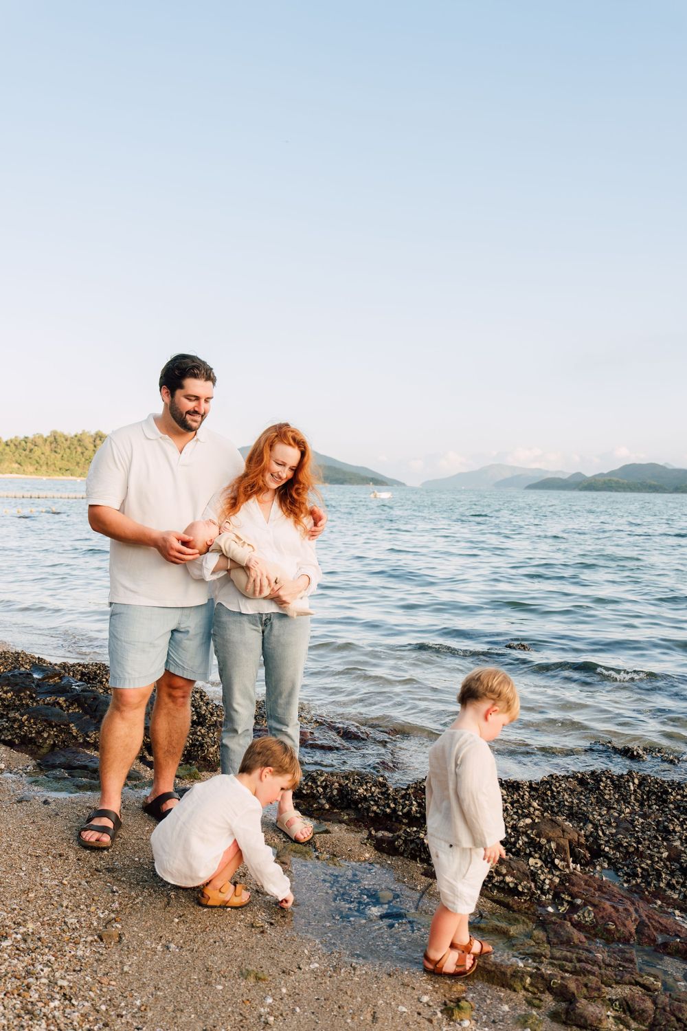 Family of four enjoying a sunny day at a rocky beach, parents holding baby while toddler plays at waters edge.