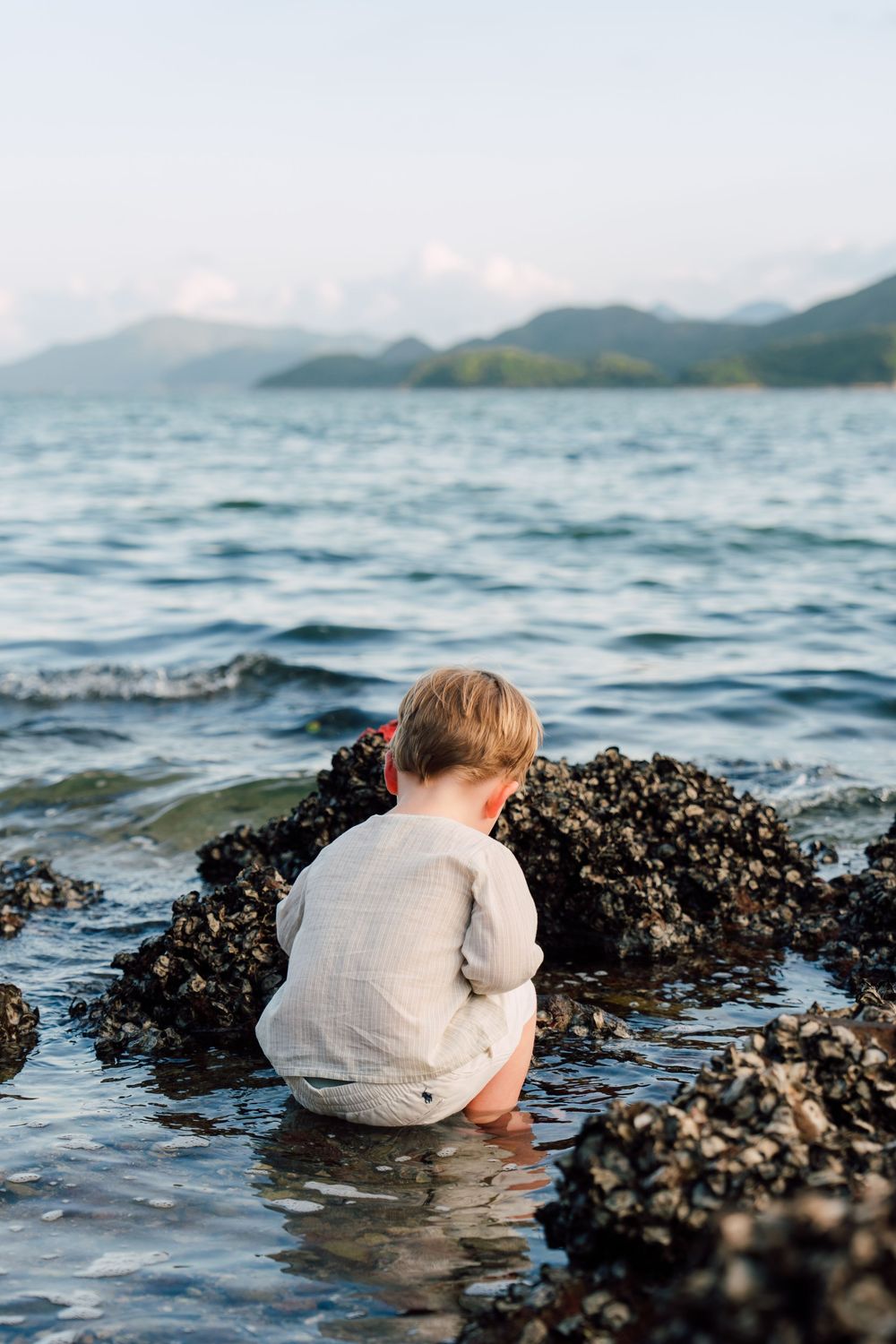 Young child sitting on rocky shore exploring tide pools with scenic ocean and mountain backdrop.