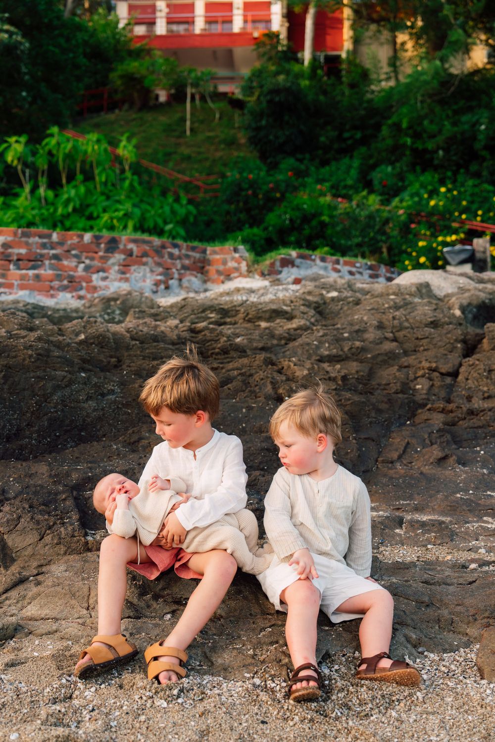 Two young boys in white linen outfits sit on rocky shore, with lush greenery and brick building behind them.