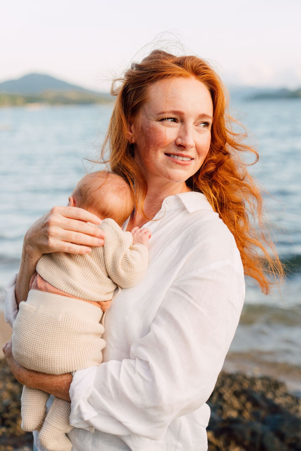 Red-haired freckled woman in white shirt holding newborn baby on rocky beach at sunset.