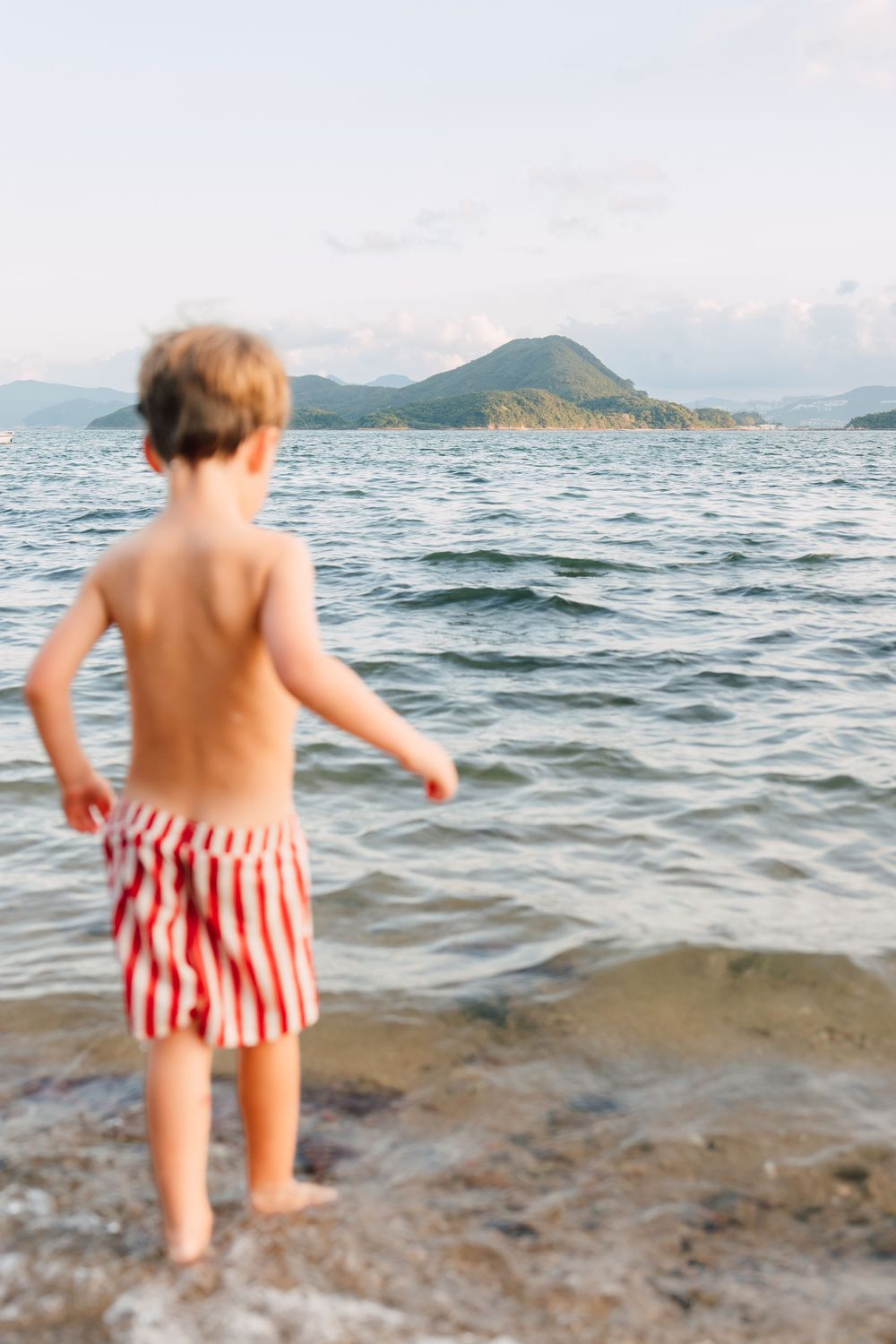 Young boy in red striped swim shorts standing at water's edge, gazing at island mountains across calm sea.