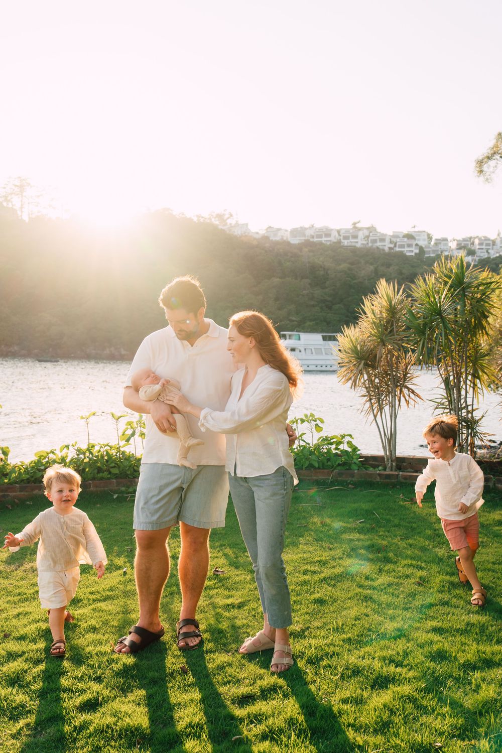 Happy family with two young children and a baby enjoying a sunny day on green lawn by a waterfront.