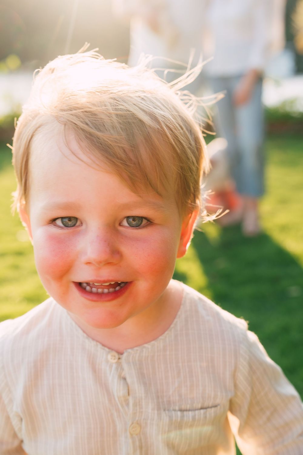 Smiling toddler with blonde hair and blue eyes outdoors in golden sunlight wearing a cream button-up shirt.