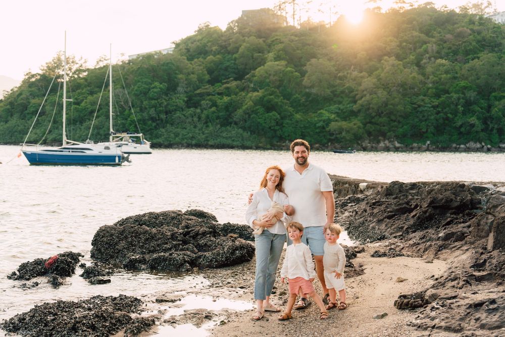 Family of four posing on a rocky beach at sunset with sailboats and lush green trees in the background.