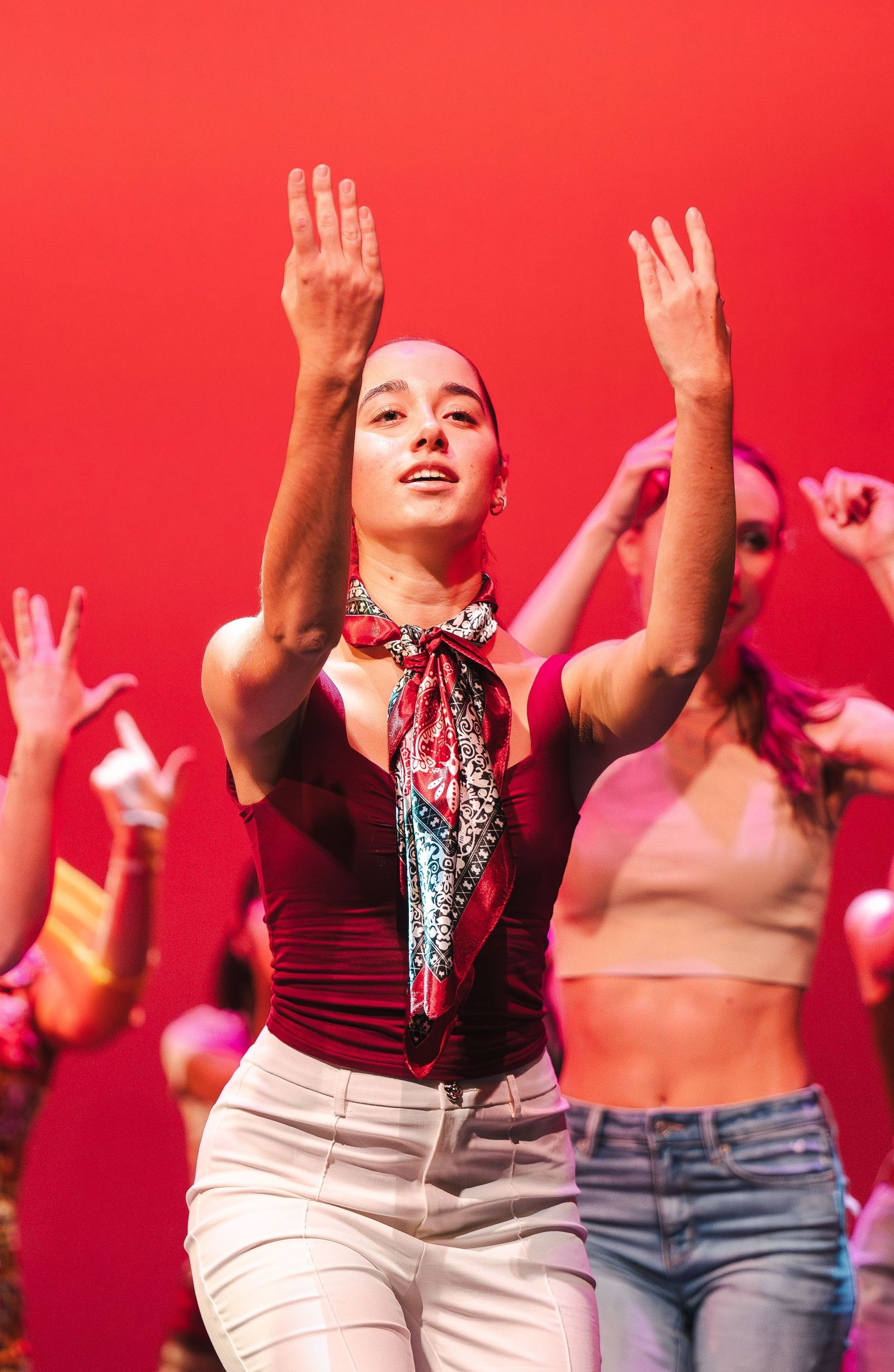 Dancers performing on stage against vibrant red backdrop with raised arms.