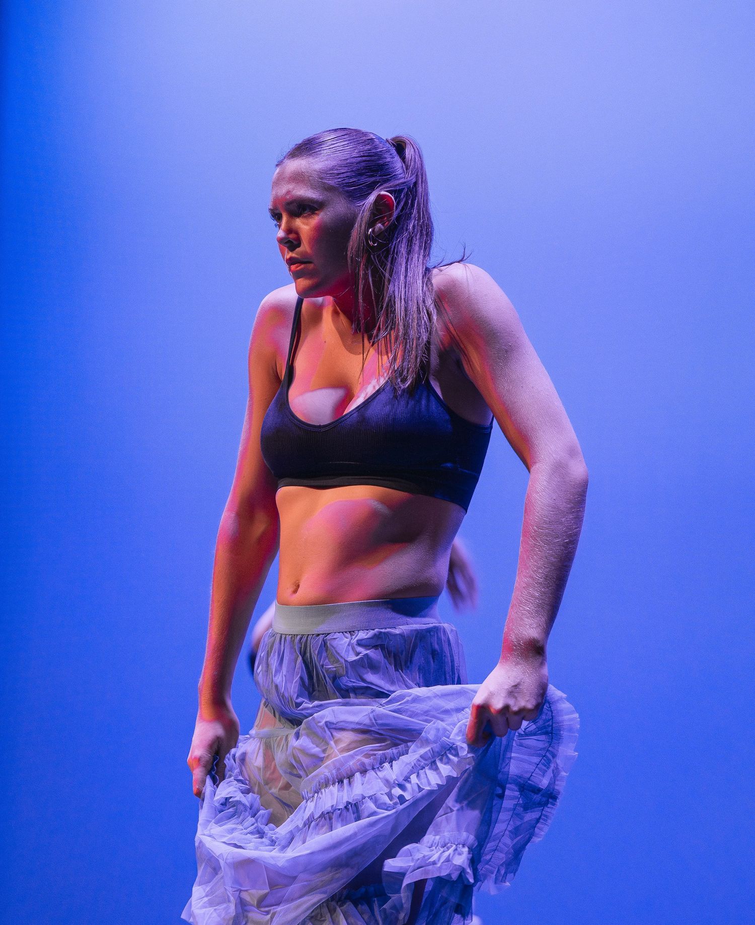 Athletic dancer in black sports bra and sheer pants performs against blue lighting on stage.