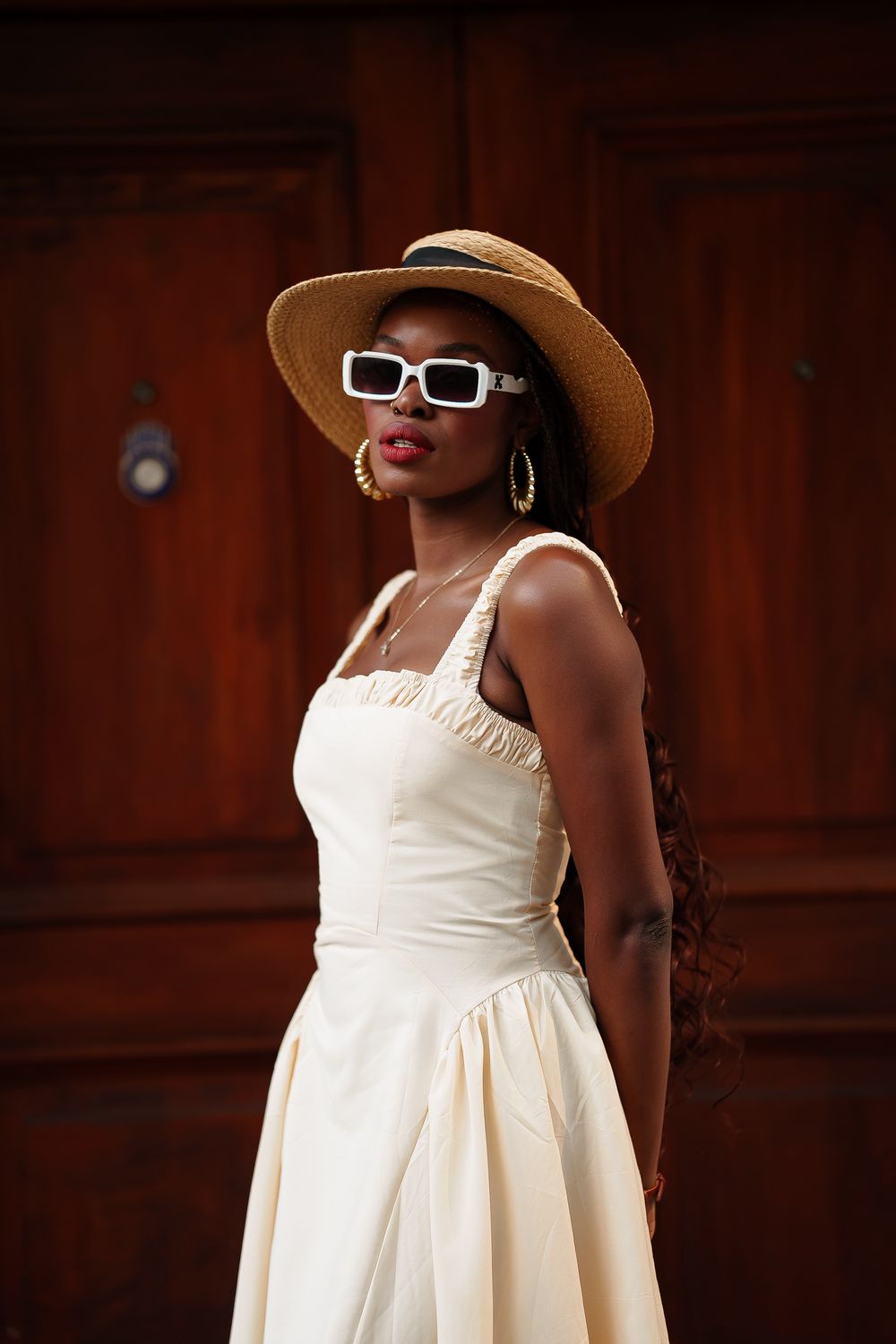 A person in a cream-colored dress and straw hat poses against a dark wooden wall.
