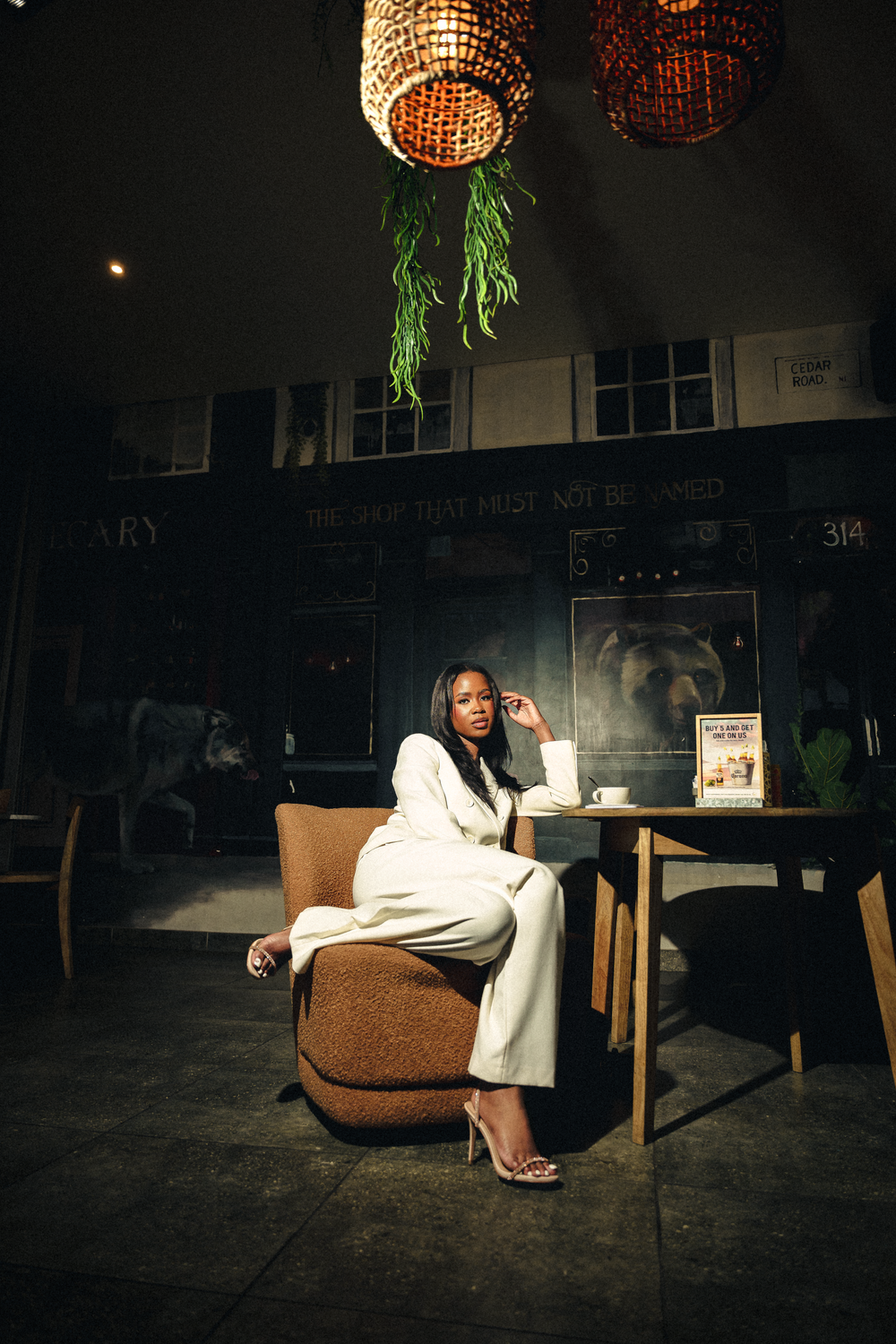 A person in white attire sits at a wooden table in a dimly lit restaurant with hanging rattan lamps and green plants.