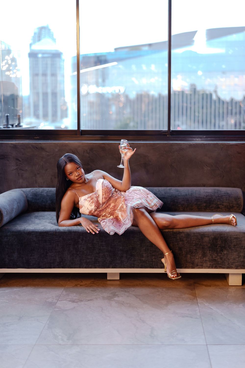 A person in a pink dress reclines across a brown leather couch near large windows in a modern space.