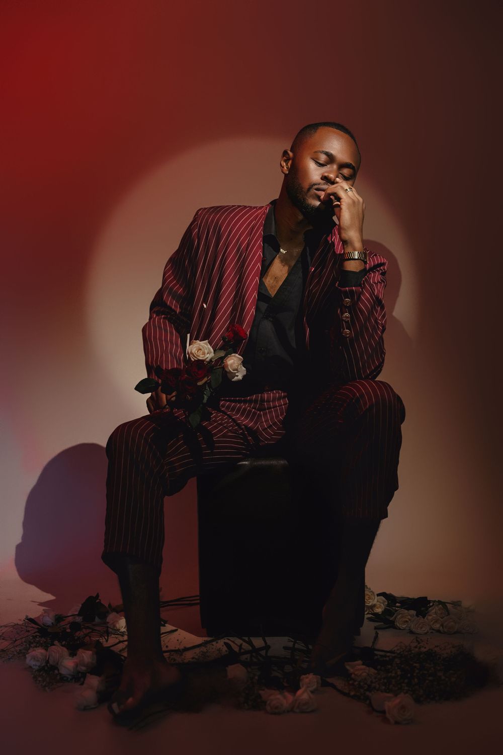 A person in a pinstriped suit sits thoughtfully on a stool against a dark red backdrop with rocks scattered on the ground.