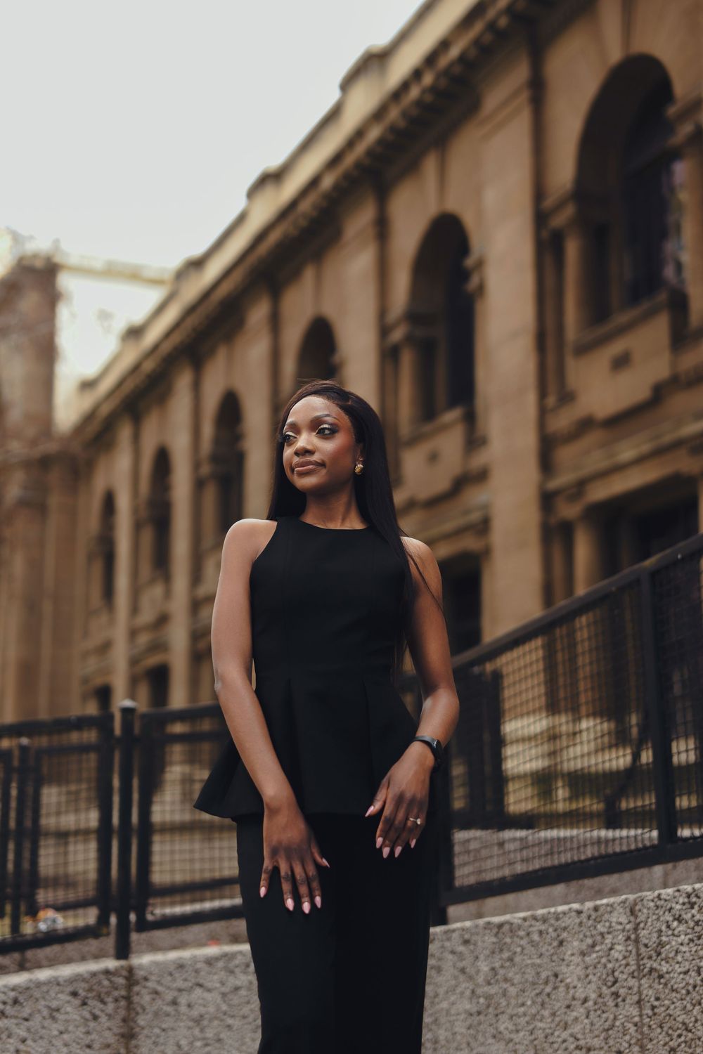 Woman in black sleeveless dress poses confidently against architectural backdrop with arched columns.