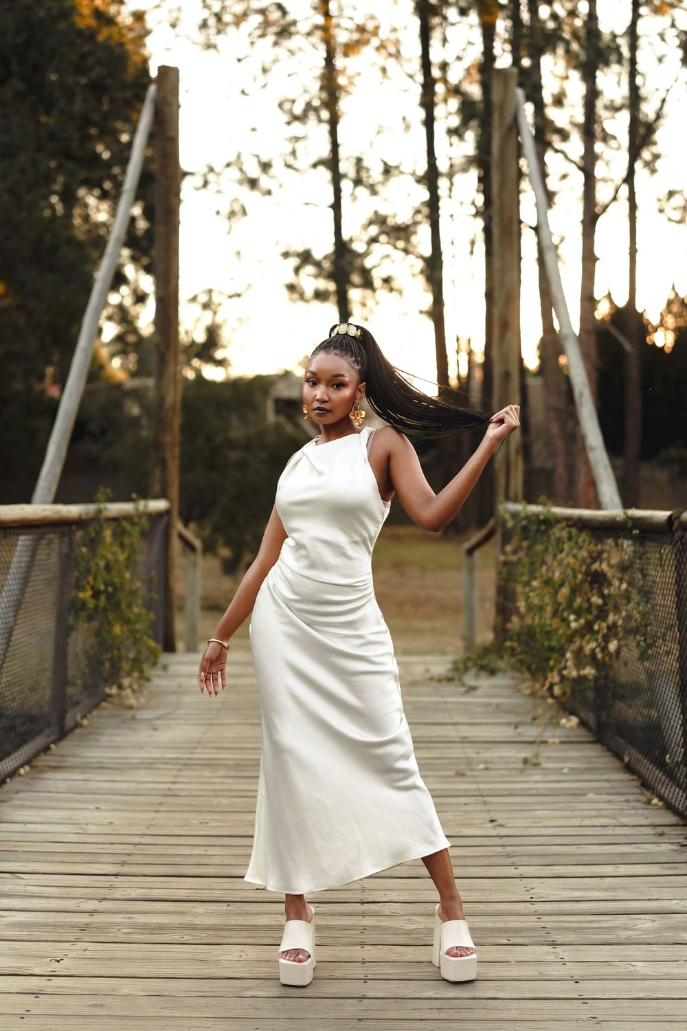 Woman in flowing white dress dances gracefully on wooden bridge surrounded by tall trees at sunset.