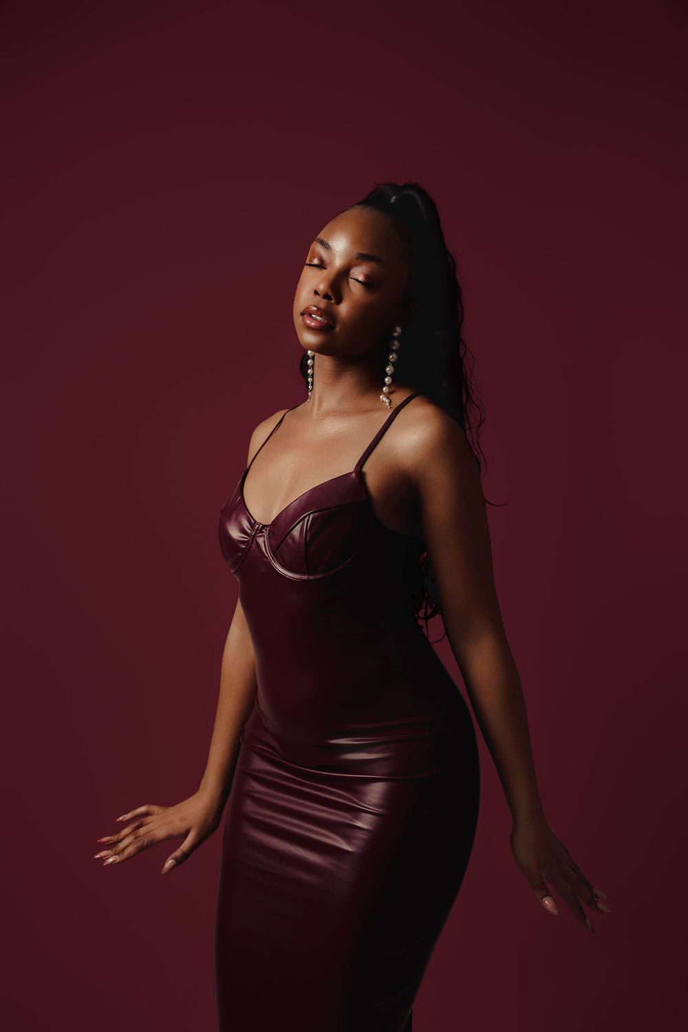 A figure poses dramatically in burgundy attire against a dark maroon background in an artistic portrait series.