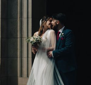 Bride and groom share a romantic moment, bride holding a bouquet in a white gown, groom in a navy suit with red tie.
