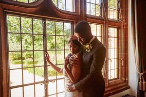 Bride and groom share a romantic embrace by ornate leaded windows in a grand historic venue.