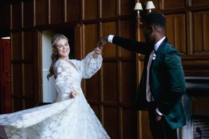 Bride in lace gown twirls as groom in green velvet suit holds her hand in elegant wood-paneled wedding venue.