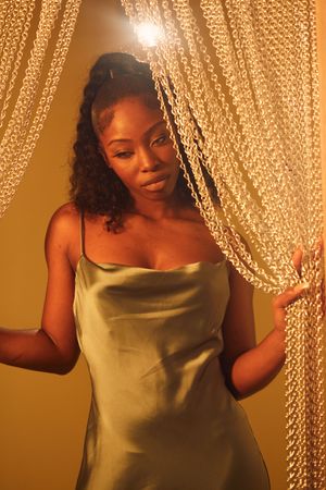 Woman in gold satin dress poses against shimmering beaded curtain backdrop in warm lighting.