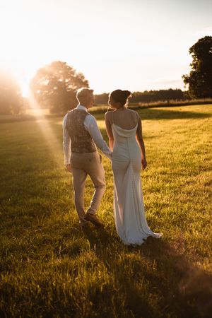 Bride and groom holding hands at golden sunset in green field, bride in elegant white gown, groom in vest and trousers.