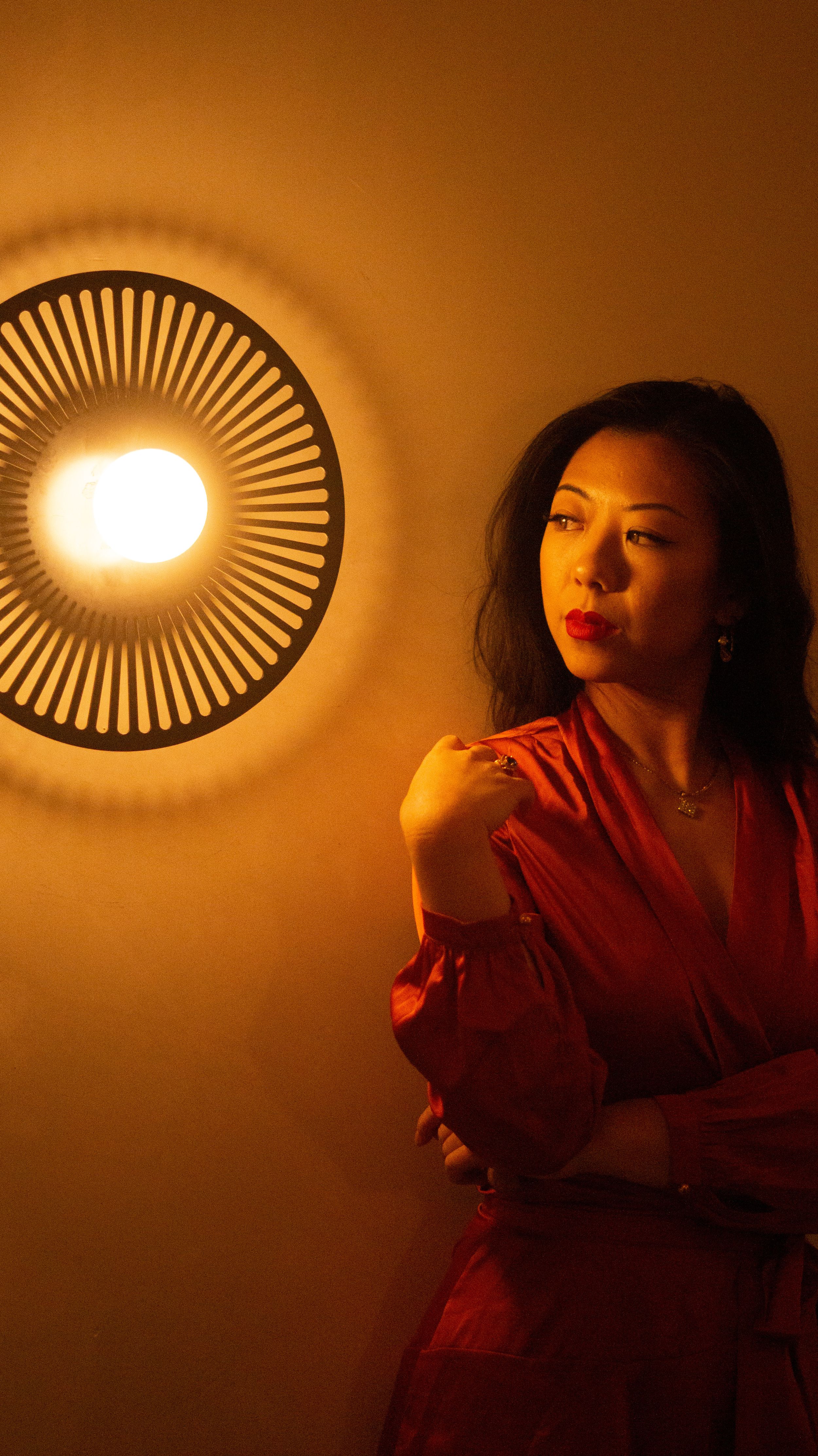 Woman in red silk dress poses by glowing circular wall sconce light in warm amber ambiance.
