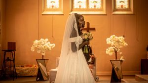 Bride in white wedding gown and veil holding bouquet at church altar with floral arrangements and stained glass windows.