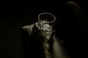 A hand holding an empty glass tumbler in dim dramatic lighting on a dark leather surface.