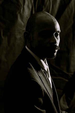 Dramatic low-key portrait of a bald Black man with mustache wearing a suit against dark fabric backdrop.