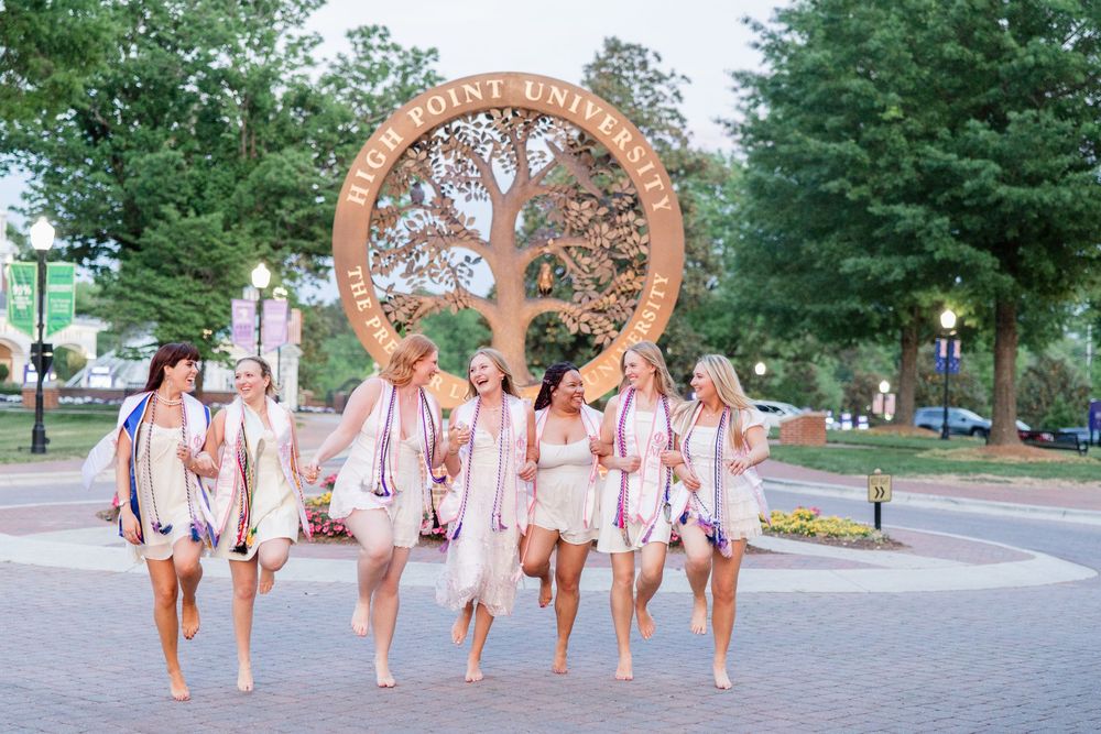 Female graduates in white dresses walk past a university seal monument on a college campus during graduation celebrations.