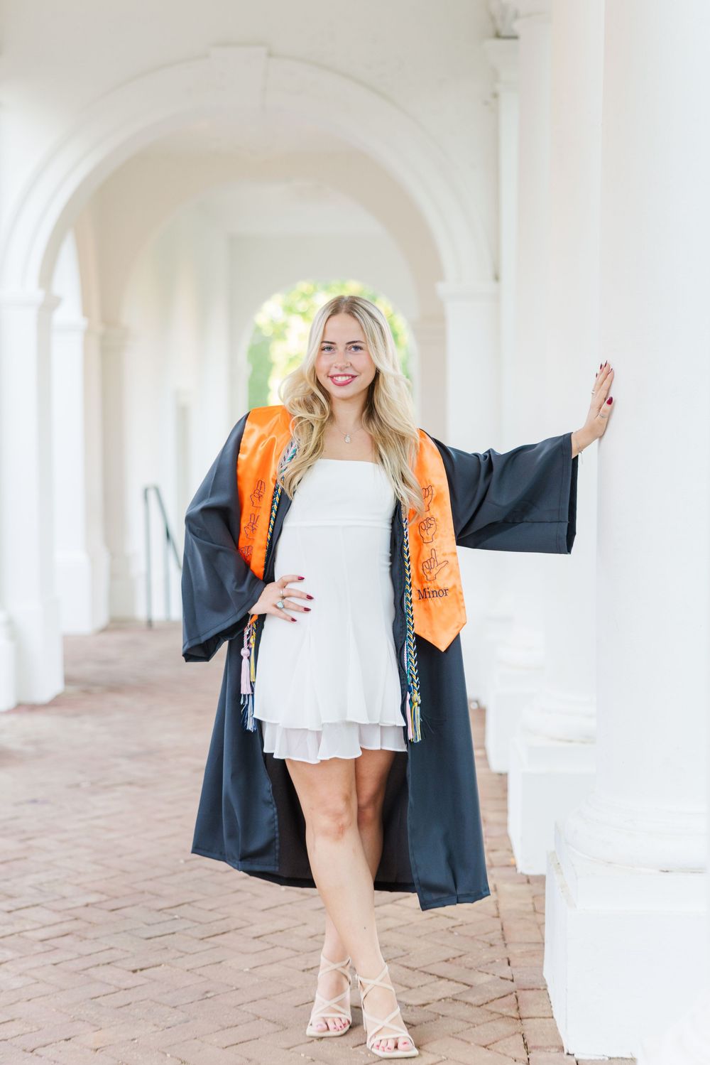 Smiling blonde graduate in black cap and gown with orange stole poses in white columned archway wearing white dress.
