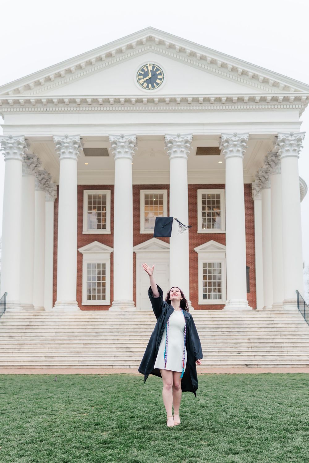 A graduate in cap and gown tosses her mortarboard in front of a classic brick and white column university building.