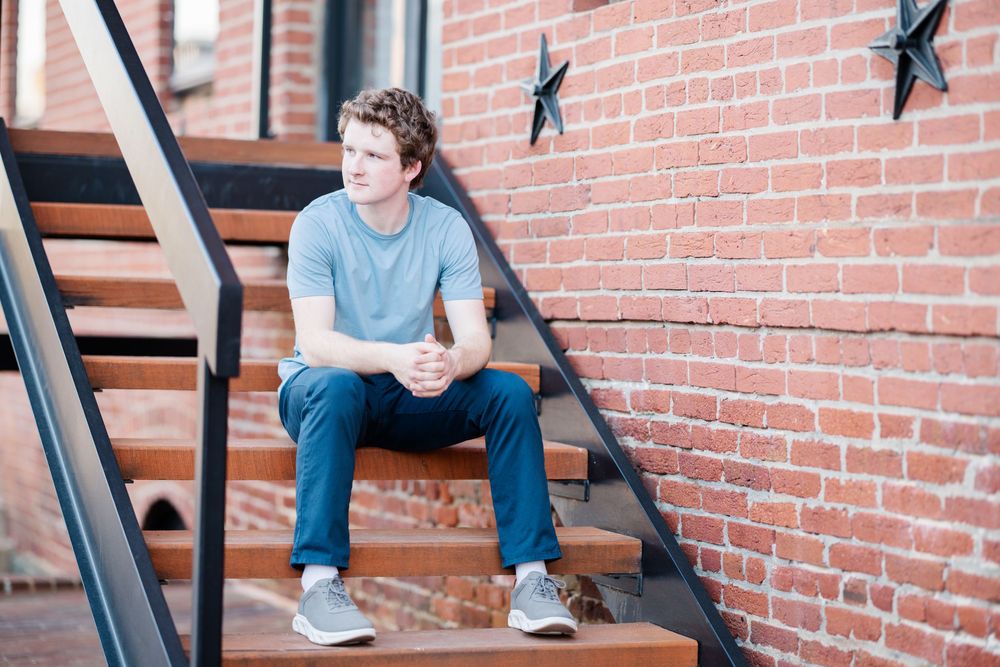 Young man in gray t-shirt and jeans sitting on wooden steps against a red brick wall with metal star decorations.