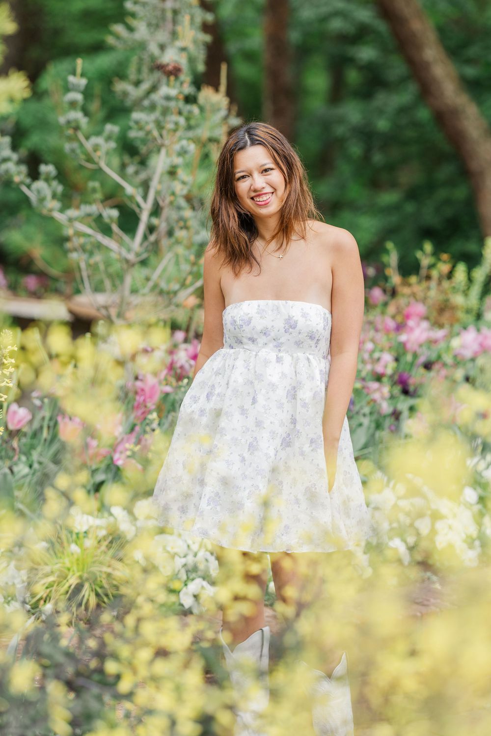 Smiling woman in strapless floral white dress standing among colorful garden flowers with lush greenery background.