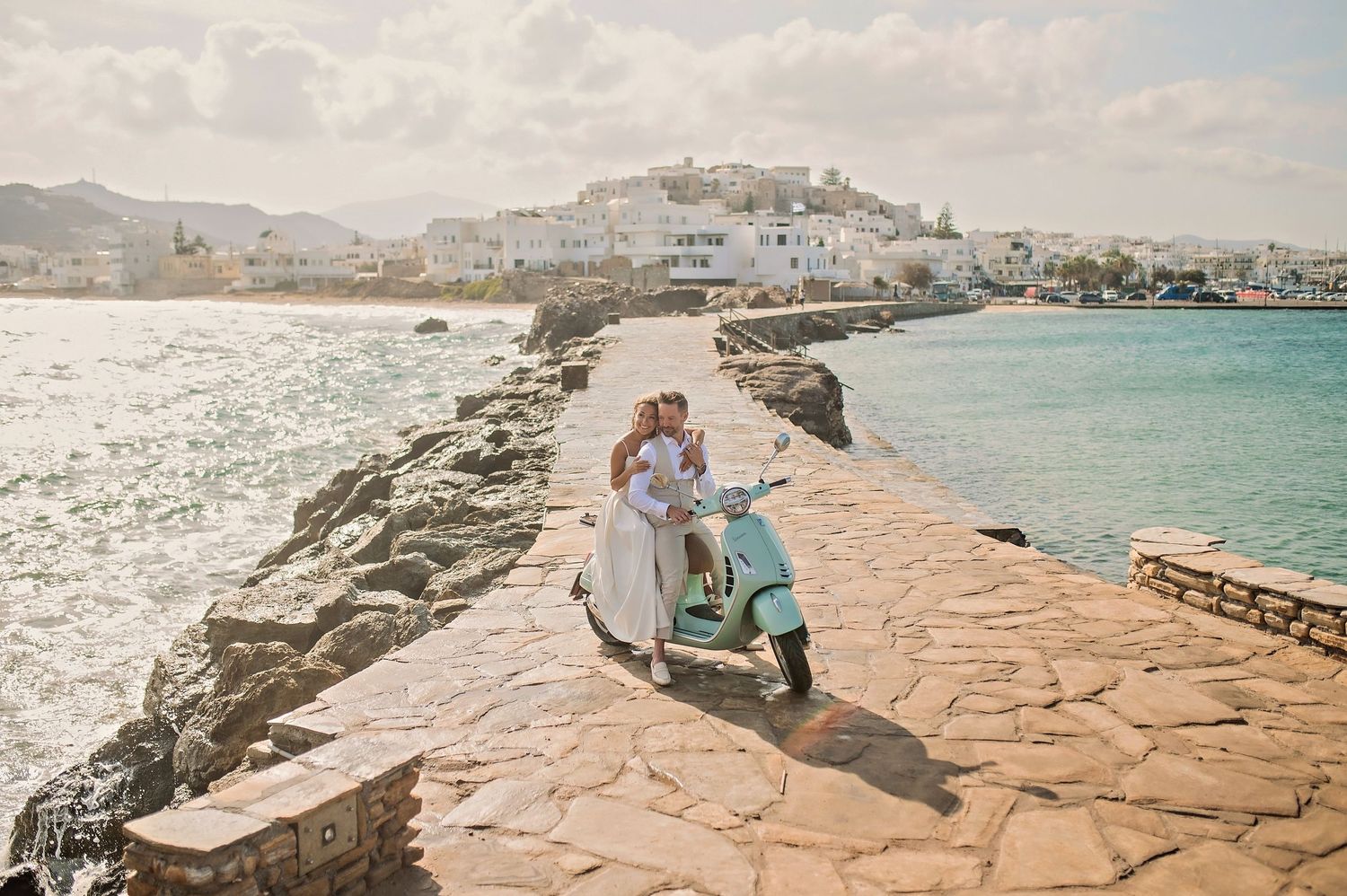 A vintage Vespa scooter parked on a stone jetty overlooking a white coastal town and turquoise waters.