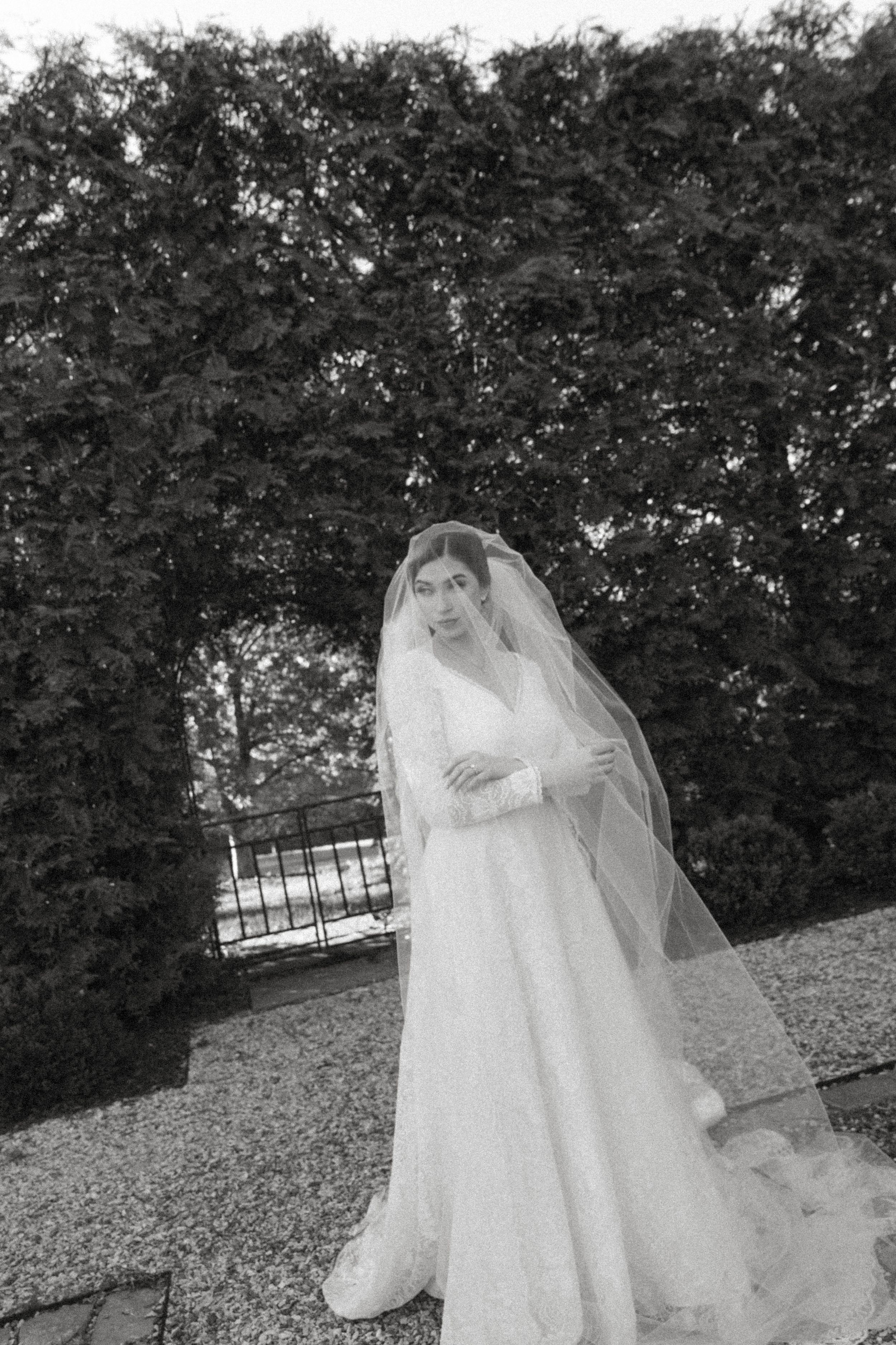 Black and white portrait of bride in long veil and wedding gown against garden backdrop.