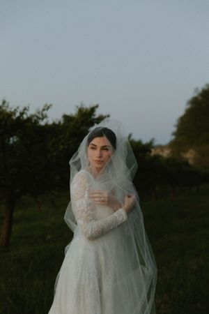 Bride in lace wedding dress and veil poses in garden during golden hour.
