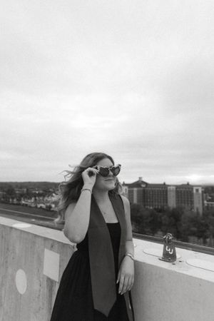 Graduate in black dress and sunglasses poses on rooftop during overcast day.