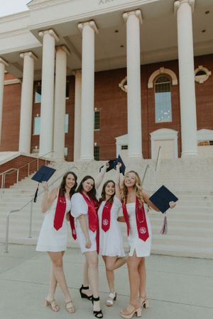 Four graduates in white dresses and red stoles celebrate in front of classical building columns.