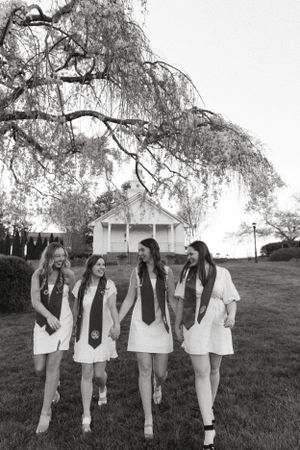 Black and white photo of four graduates walking together under willow tree branches.