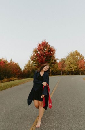 Graduate in a black dress and coat walks barefoot on a road with autumn trees in the background.