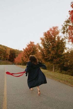 Person in dark coat dances on autumn road with red scarf flowing in wind.