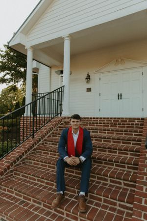 Graduate in navy blazer and red tie sits on brick steps of white columned building.