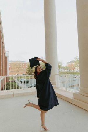 Graduate in cap and gown twirls joyfully on a columned porch during graduation celebrations.