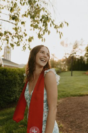 A graduate in white dress with red sash laughs in sunlit garden setting.