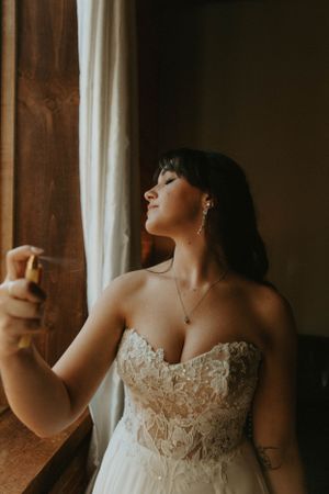 A bride in lace wedding dress poses by window in soft natural light.