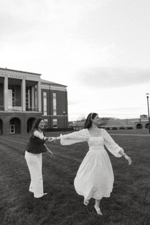 Two people dancing joyfully on grass in front of a brick building in black and white.