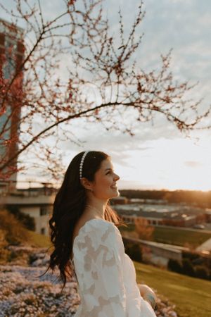 A person in white dress with pearl headband poses against sunset and cherry blossoms.