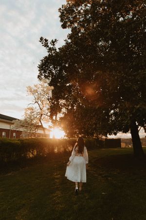 A person in white dress stands beneath a tree during golden hour sunset.